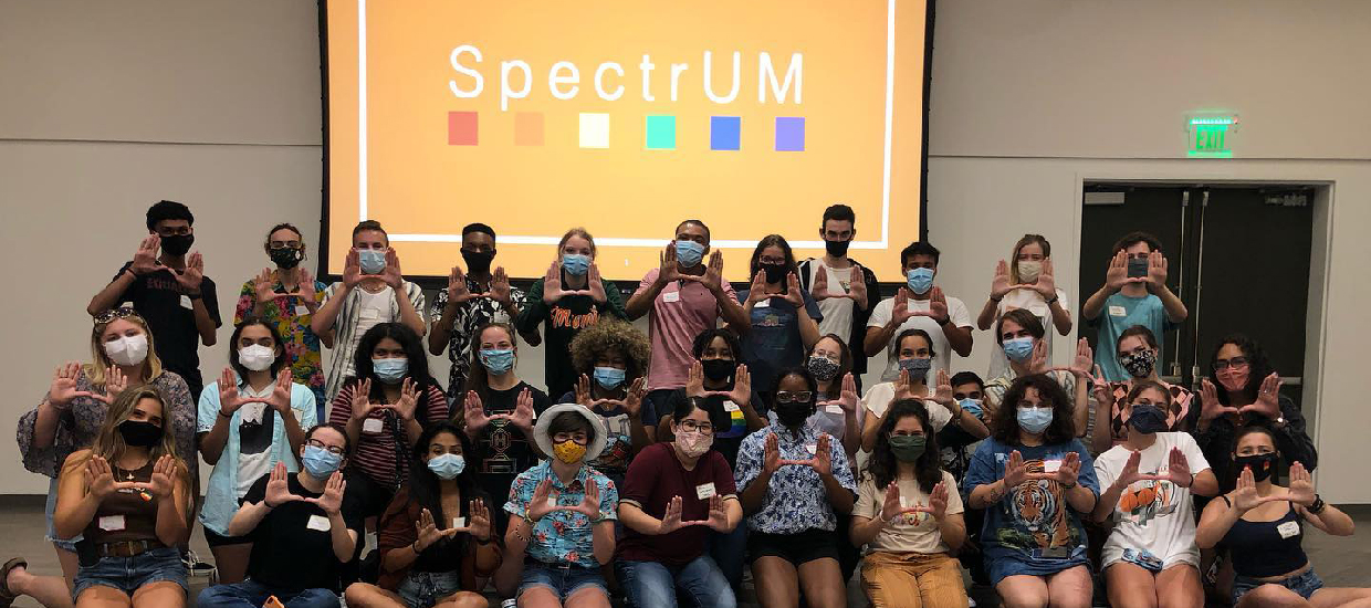A group of SpectrUM students at an event stand together, holding up the U sign with their hands. They are all wearing face masks. 